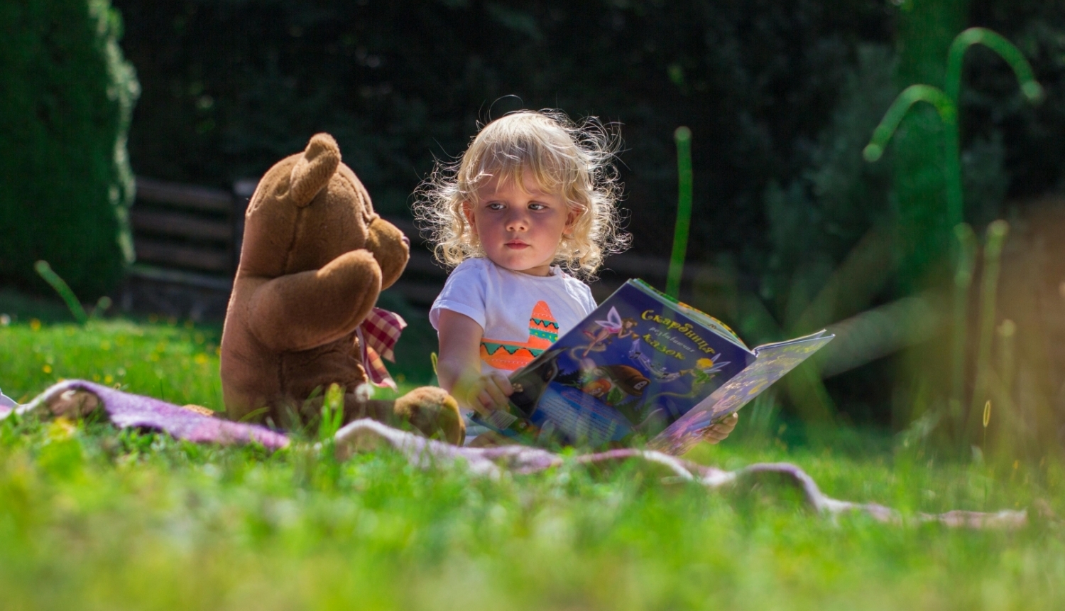 Ilustratīvs attēls - bērns sēž pļavā ar lācīša rotaļlietu 